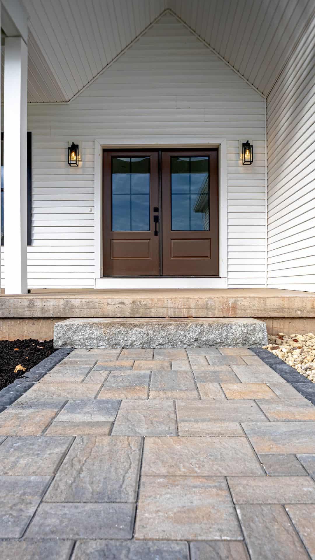 Edited camera image of sidewalk completed Front porch of a white house featuring double brown doors, a stone step, and a newly installed paver walkway with decorative landscaping and river rocks.